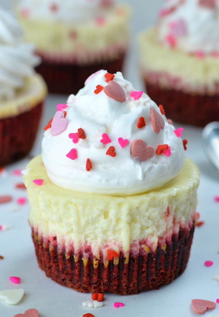 Red velvet cheesecake cupcakes topped with whipped cream and garnished with festive heart shape sprinkles