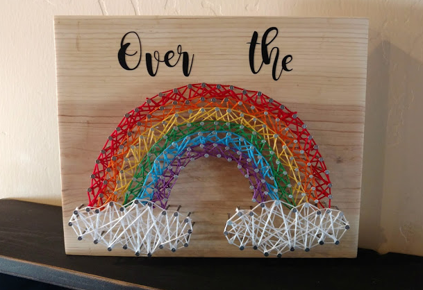 Rainbow string art above a rectangle piece of wood that has a text saying over the