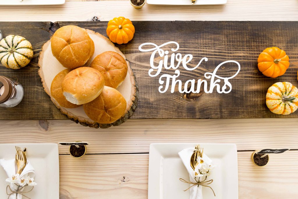 Wooden table runner with text on it saying give thanks, and some breads and pumpkins on top of it