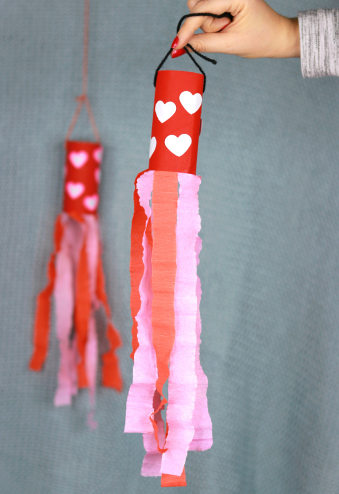 Valentine's day windsock made from a toilet paper roll painted red with heart shapes on it and a pink and red tissue paper strips on it.
