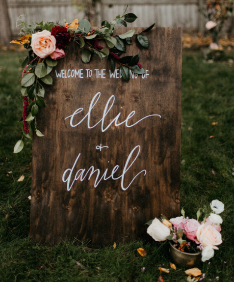 Calligraphy wedding sign on a wood that says Welcome to the wedding of Ellie & Daniel with flower arrangement on it