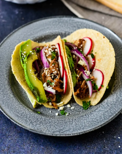 slow cooker chocolate mole chicken tacos serve with cotija cheese, chopped cilantro, sliced avocado, lime sections, sliced radishes and red onions.
